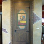 Pantry Door painted to look like old wood with old wrought iron hardware, beautiful signage with a rooster. Walls painted to look like old stucco, cracked and broken away to reveal bricks. Stone surround on the doorframe. Mural by Ellen Leigh