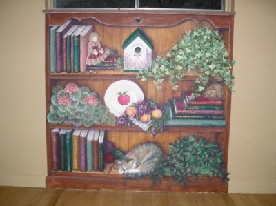 Book case mural on a wall below a window to look built in. Filled with favorite book titles and favorite pastimes of the family. Portrait of a favorite cat. Mural by Ellen Leigh